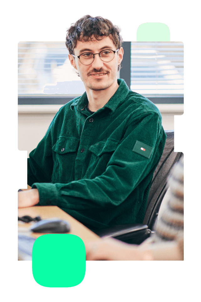 A man with short curly brown hair, glasses, and a green corduroy shirt is seated at a desk, looking to his right and smiling slightly. He is in an office setting with a window and blinds in the background. Another person is partially visible in the foreground.